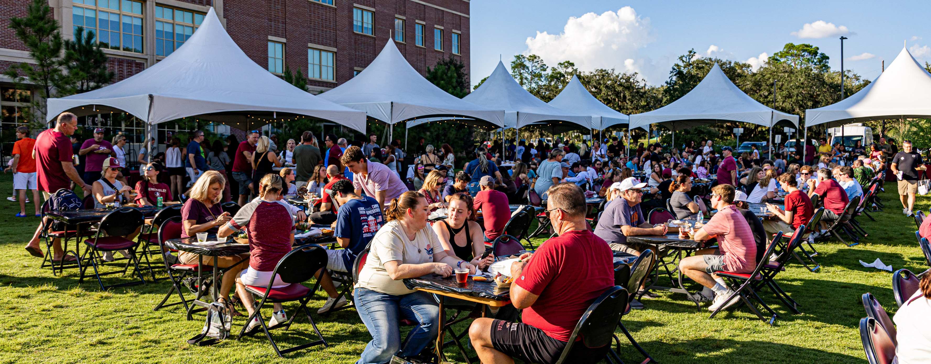 Large outdoor gathering at a university campus with many families and students seated at round tables on a lawn, eating and talking under white event tents beside a brick building on a sunny day.