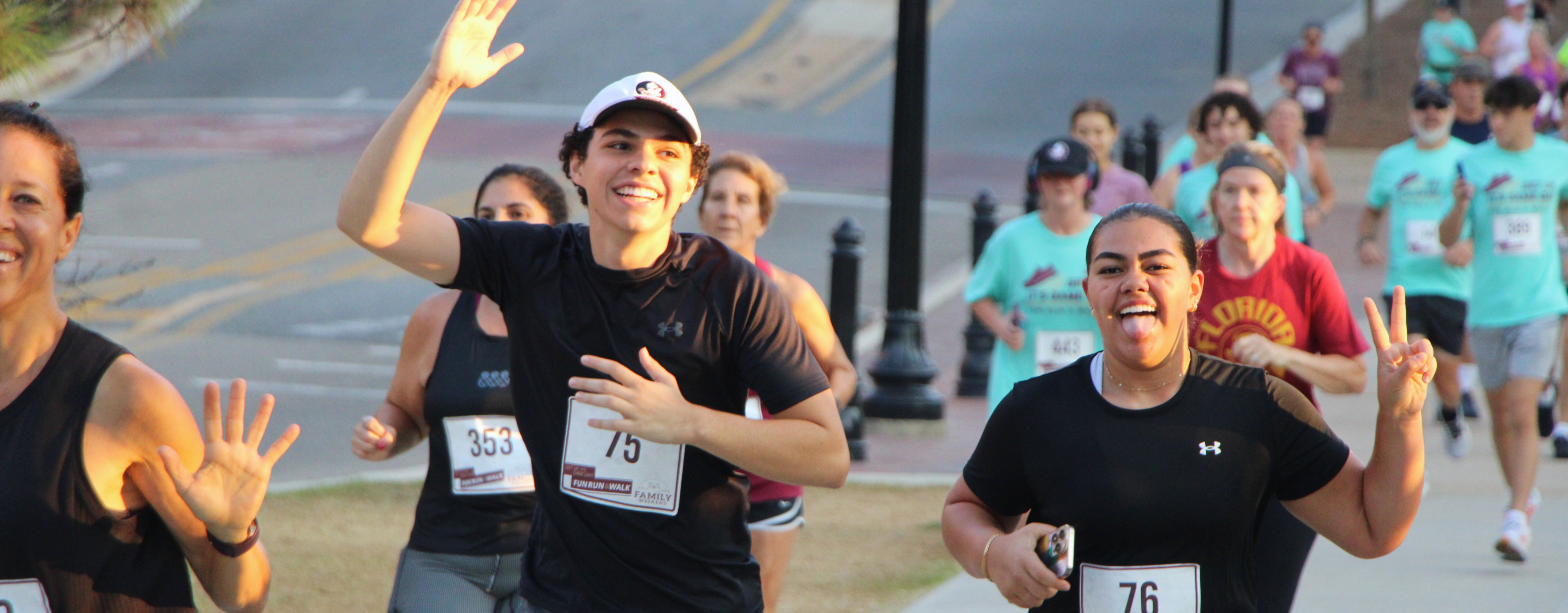 Participants in a campus fun run jog along a street, smiling and waving at the camera; one runner raises a hand while another flashes a peace sign, with a group of runners following behind.