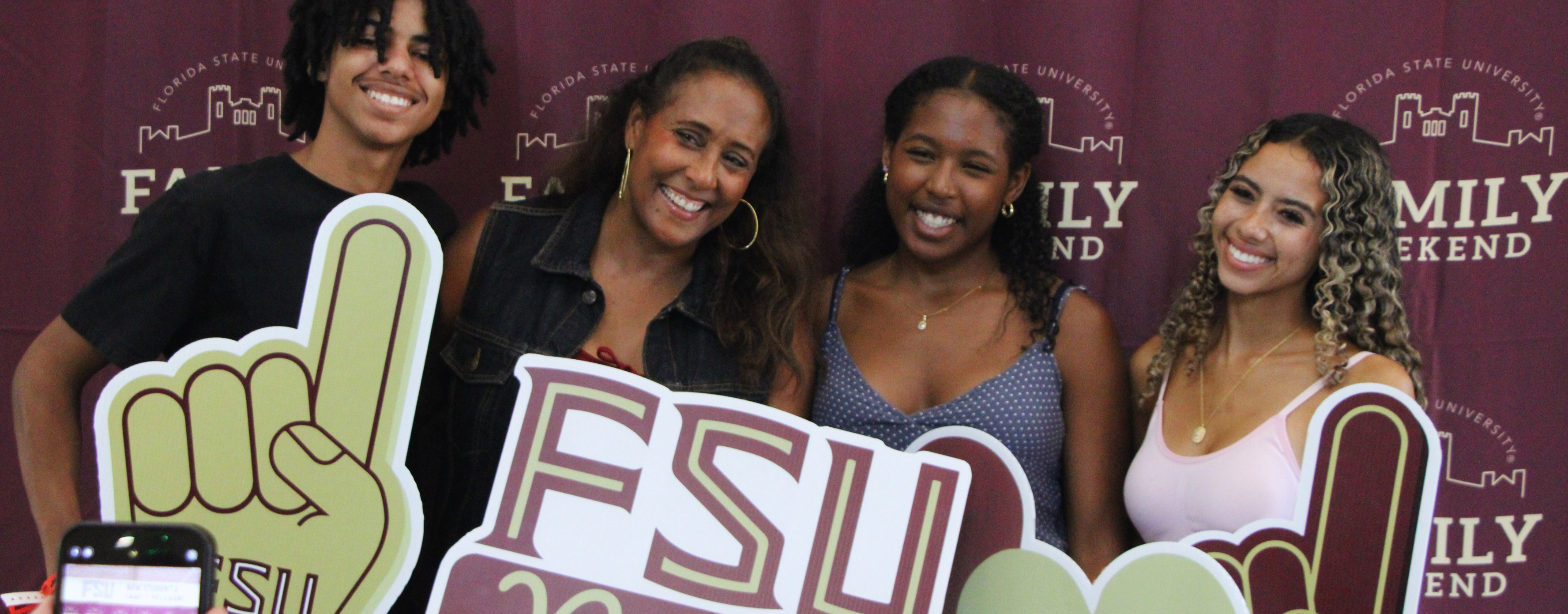 Four smiling people pose in front of a Florida State University “Family Weekend” backdrop, holding large FSU-themed props including foam fingers and a sign that reads “FSU.”