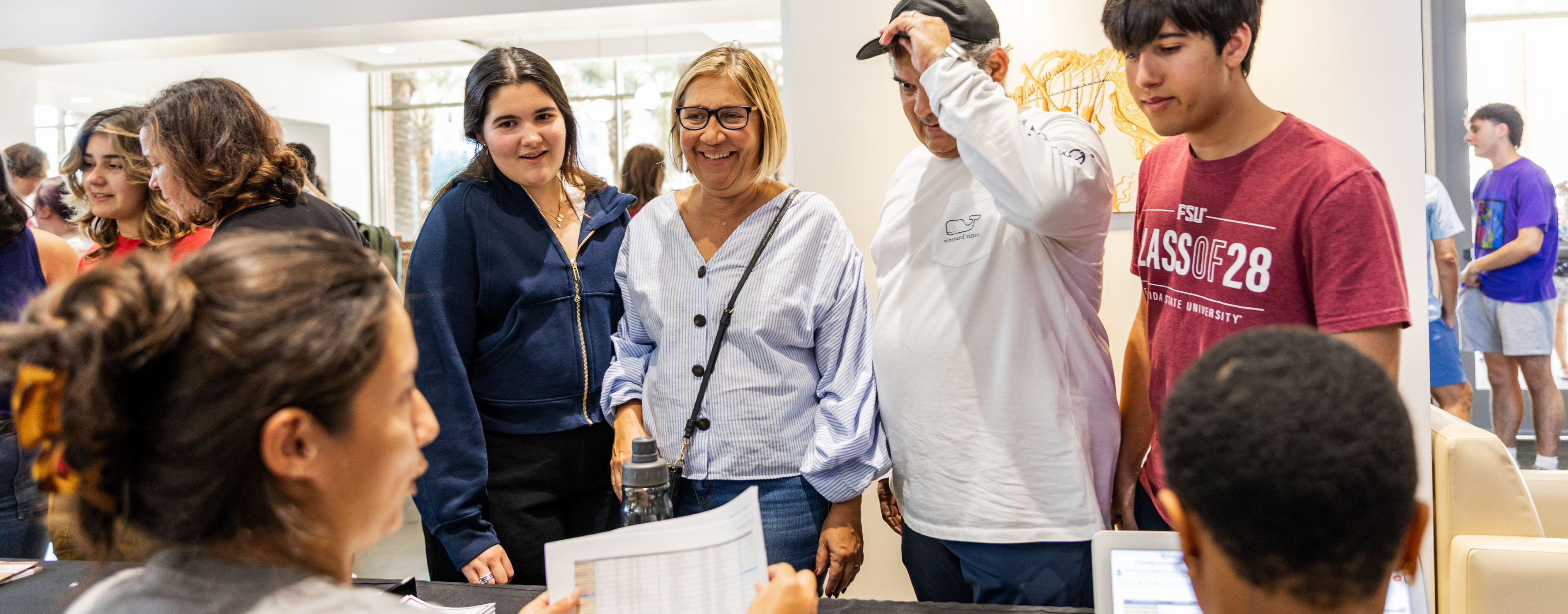 A group of people, including a smiling woman in glasses, stand together indoors, appearing engaged and happy.
