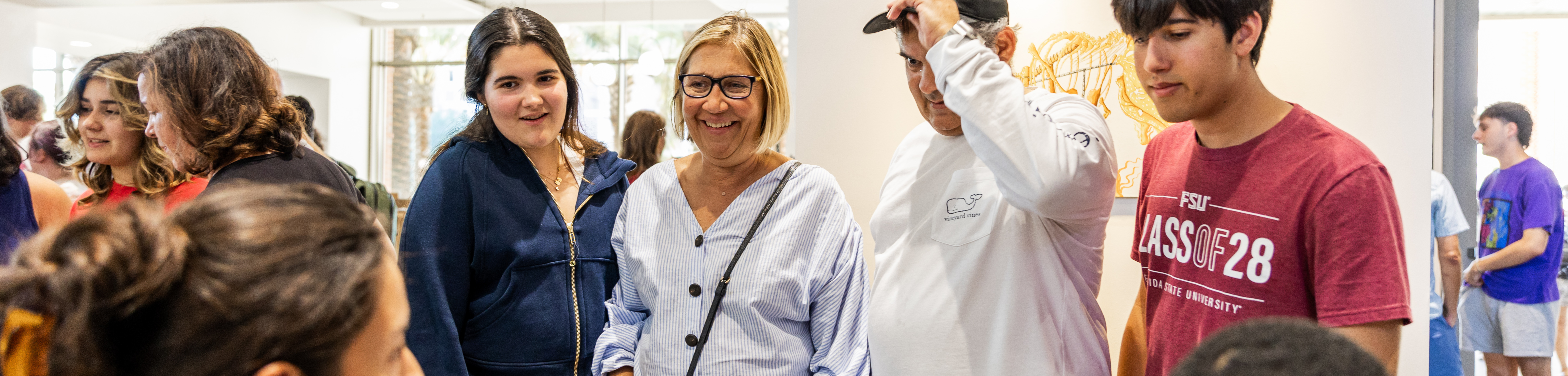 A group of people, including a smiling woman in glasses, stand together indoors, appearing engaged and happy.