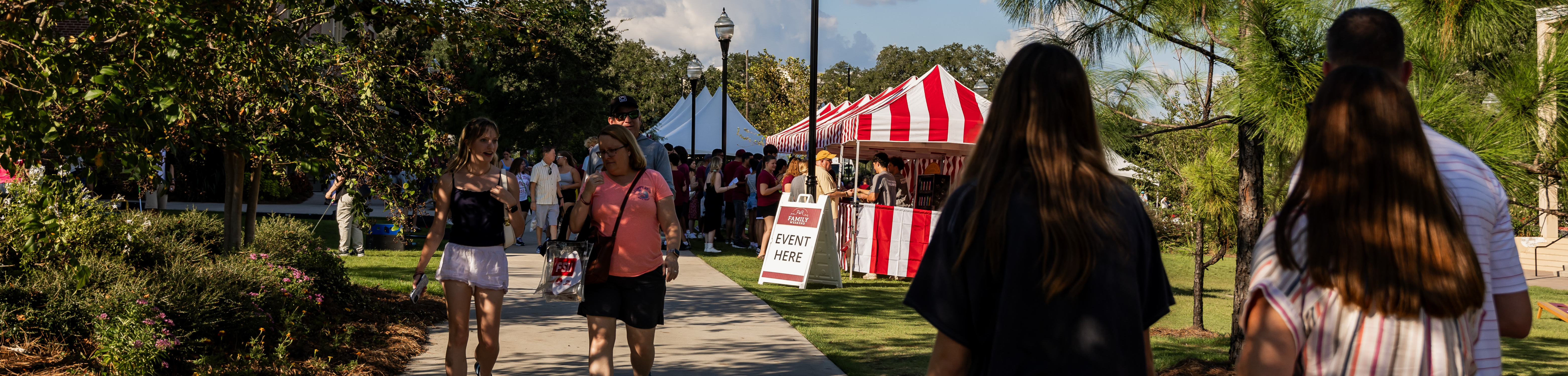 People are walking along a path at an outdoor event with a red and white striped tent nearby.